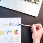 Hand holding pencil reviewing colorful data charts on desk with laptop.